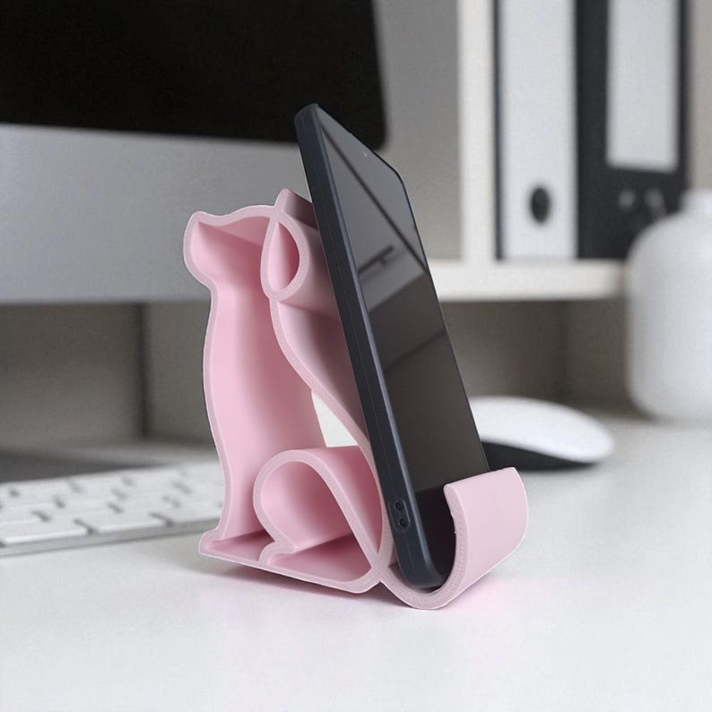 May include: A light pink phone stand shaped like a dog, holding a black smartphone. The stand has a curved design and is made of a smooth material. The phone is positioned vertically, displaying a dark screen. The stand is on a white surface.