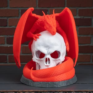 May include: A white skull sculpture with a red dragon perched on top and a red snake coiled around the base. The dragon has outstretched wings. The sculpture is on a dark gray base.