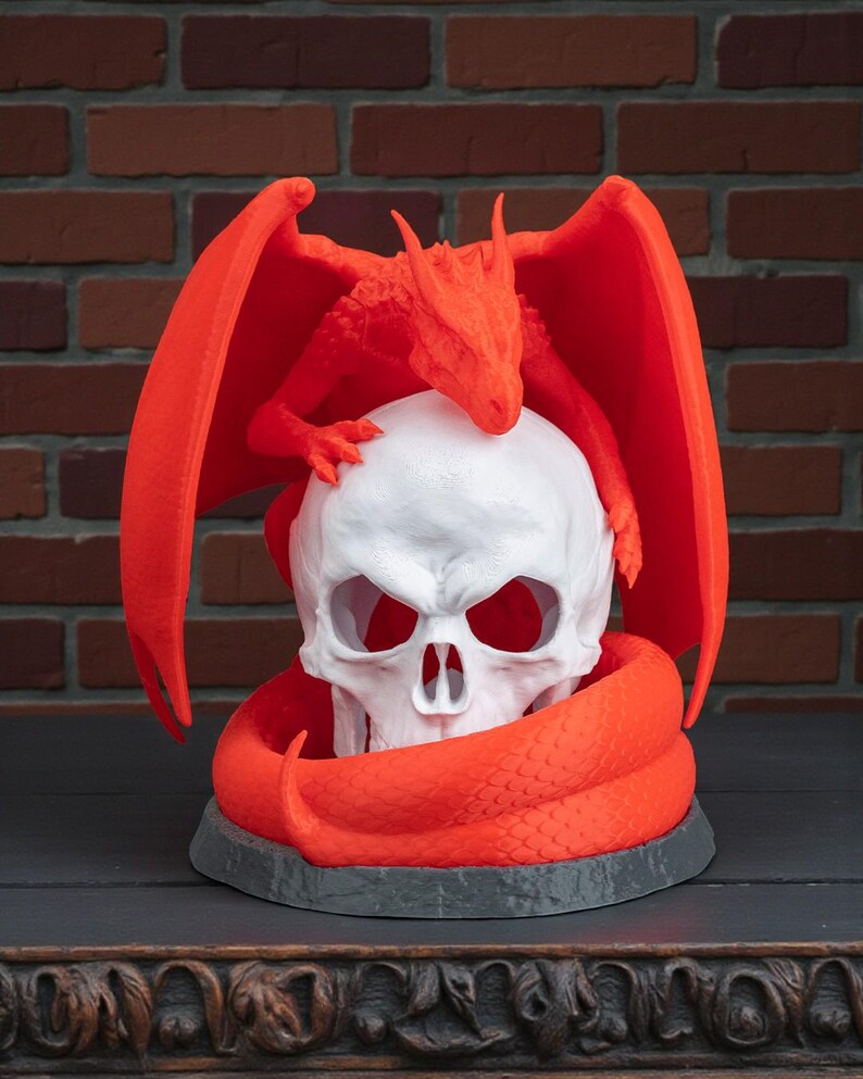 May include: A white skull sculpture with a red dragon perched on top and a red snake coiled around the base. The dragon has outstretched wings. The sculpture is on a dark gray base.