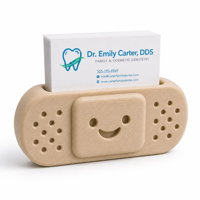 Op de afbeelding: Een lichtgekleurde houten visitekaarthouder in de vorm van een pleister. De houder heeft een lachend gezicht en bevat een stapel witte visitekaartjes met de tekst "Dr. Emily Carter, DDS".