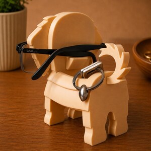 May include: A tan-colored, geometric lion-shaped eyeglasses and ring holder. The lion has a mane, tail, and legs. Black eyeglasses rest on the lion's head, and a silver ring hangs from the lion's body. The holder sits on a wooden surface.