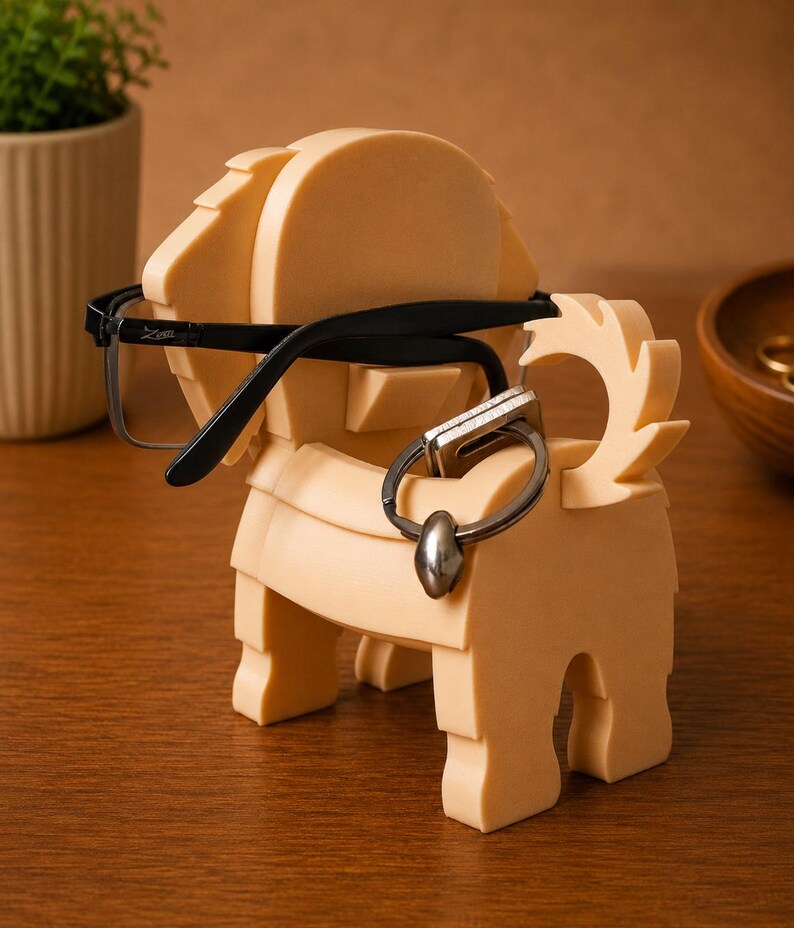May include: A tan-colored, geometric lion-shaped eyeglasses and ring holder. The lion has a mane, tail, and legs. Black eyeglasses rest on the lion's head, and a silver ring hangs from the lion's body. The holder sits on a wooden surface.