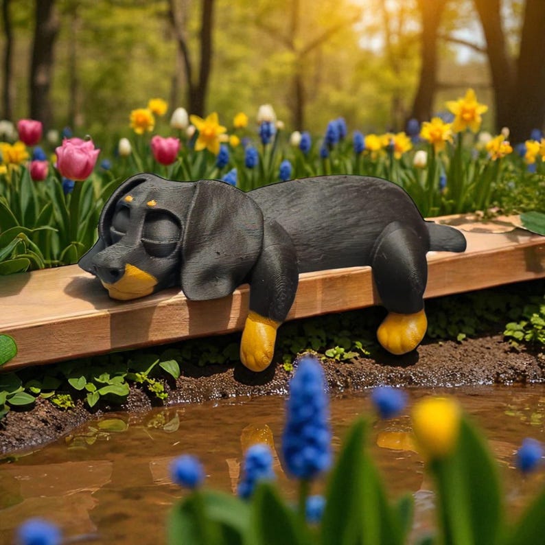 Pu&ograve; includere: Una statuina di cane bassotto nero e giallo appoggiata su una superficie di legno. Il cane ha gli occhi chiusi e dettagli gialli sulle zampe e sul muso. La statuina &egrave; ambientata in un giardino fiorito.