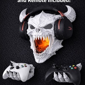 May include: A white skull-shaped wall decor with horns, wearing black and red headphones. The skull's mouth is open, revealing an orange glow. Two game controllers, one white and one black, are held by skeletal hands. Text at the top reads "RGB Puck Light and Remote Included!"