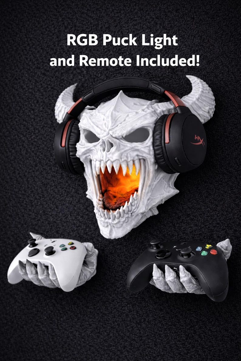May include: A white skull-shaped wall decor with horns, wearing black and red headphones. The skull's mouth is open, revealing an orange glow. Two game controllers, one white and one black, are held by skeletal hands. Text at the top reads "RGB Puck Light and Remote Included!"