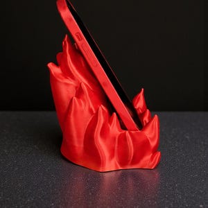 Puede incluir: Un tel&eacute;fono rojo se coloca en un soporte para tel&eacute;fono rojo. El soporte tiene un dise&ntilde;o en forma de llama. El tel&eacute;fono y el soporte est&aacute;n sobre una superficie oscura con un fondo negro.