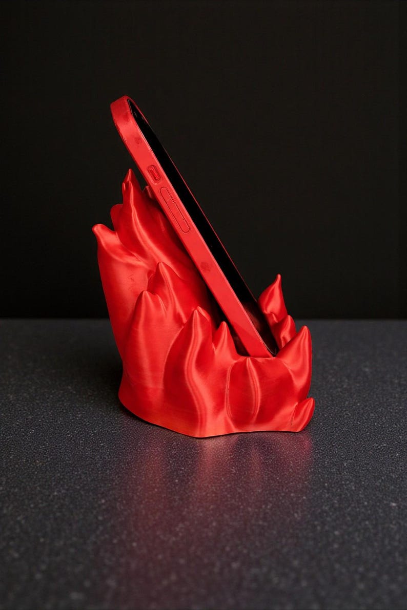 Puede incluir: Un tel&eacute;fono rojo se coloca en un soporte para tel&eacute;fono rojo. El soporte tiene un dise&ntilde;o en forma de llama. El tel&eacute;fono y el soporte est&aacute;n sobre una superficie oscura con un fondo negro.