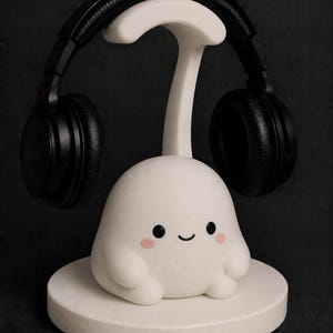 May include: A white headphone stand featuring a cute, cartoonish character with a smiling face and pink cheeks. Black headphones rest on the stand. The stand is on a round base, all in white, against a dark background.