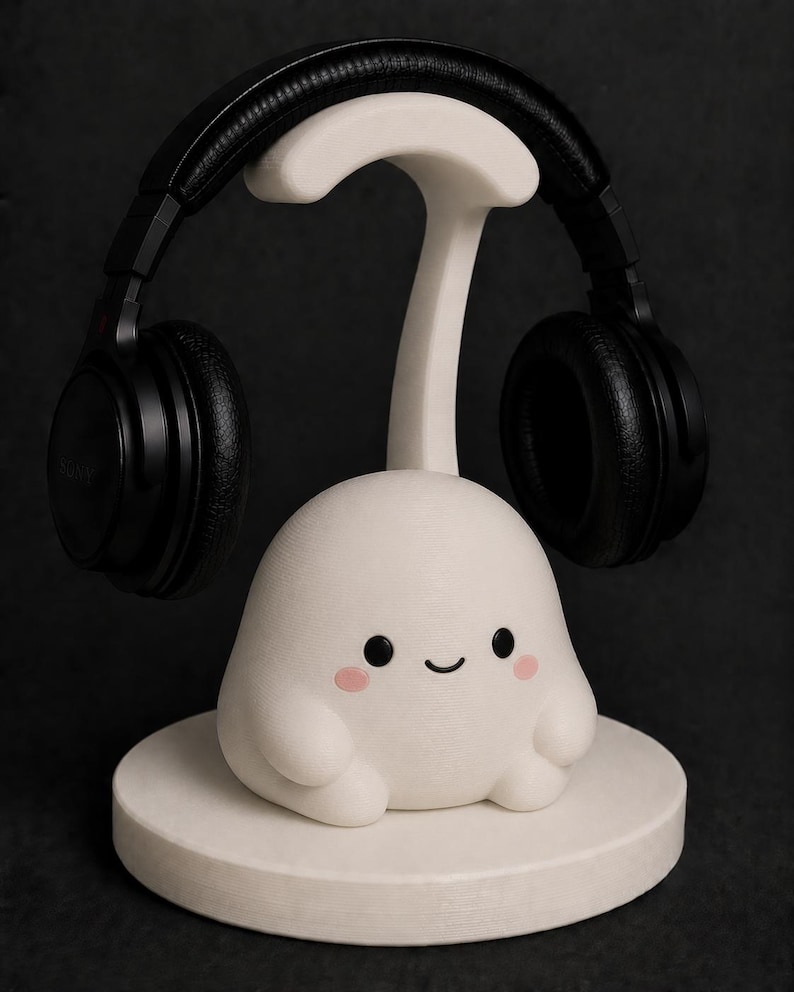 May include: A white headphone stand featuring a cute, cartoonish character with a smiling face and pink cheeks. Black headphones rest on the stand. The stand is on a round base, all in white, against a dark background.