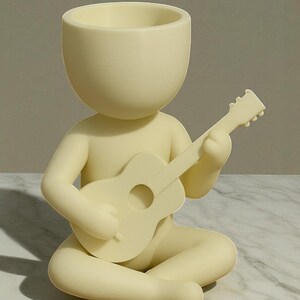 May include: A cream-colored figurine of a person seated cross-legged, playing a guitar. The figure has a large, open-topped head, functioning as a planter. The guitar is detailed with a sound hole and frets. The figurine is made of a smooth, matte material.