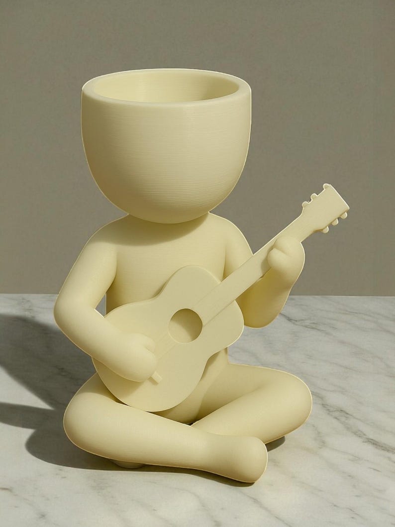 May include: A cream-colored figurine of a person seated cross-legged, playing a guitar. The figure has a large, open-topped head, functioning as a planter. The guitar is detailed with a sound hole and frets. The figurine is made of a smooth, matte material.