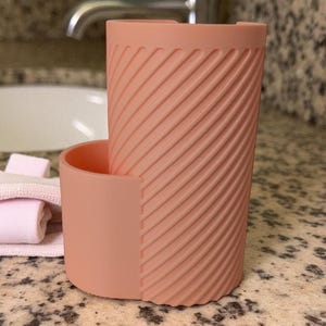 May include: A peach-colored, cylindrical toothbrush holder with a textured, diagonal stripe design. The holder has a separate, attached compartment. The holder is sitting on a speckled countertop near a sink and faucet.