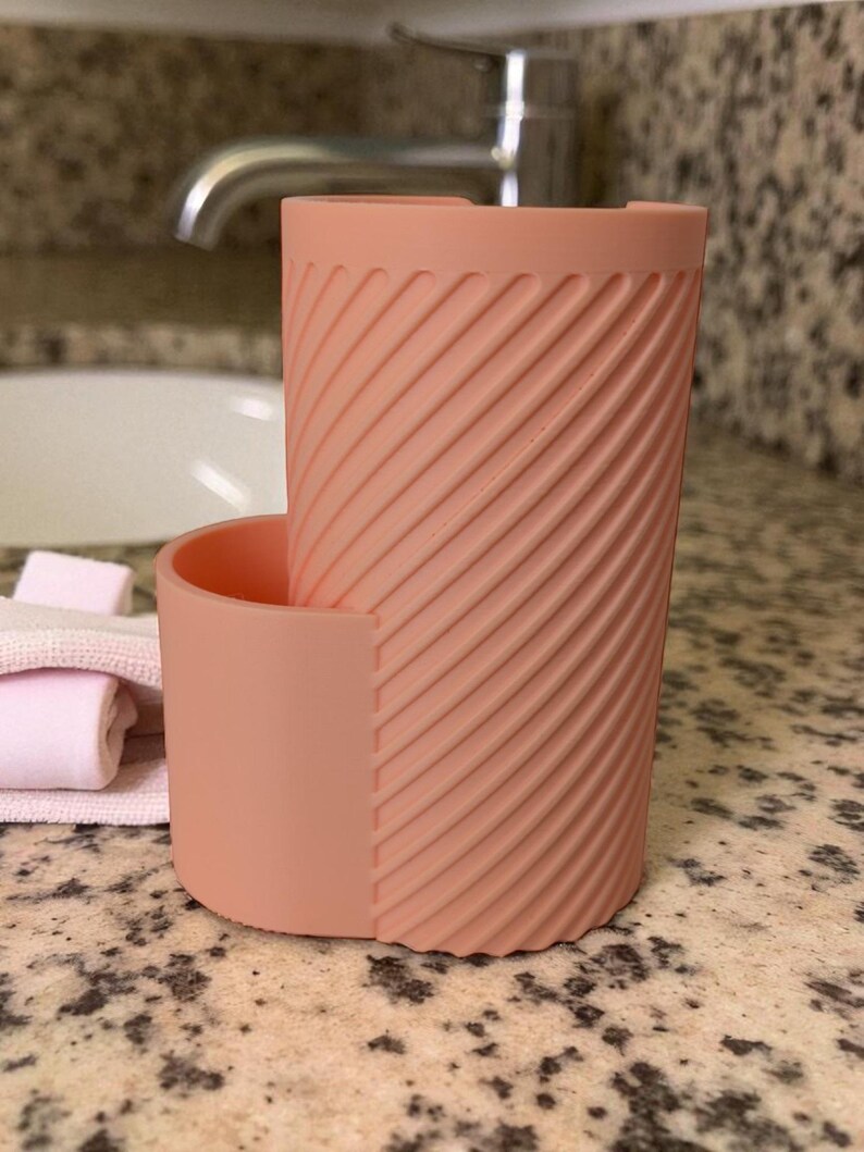 May include: A peach-colored, cylindrical toothbrush holder with a textured, diagonal stripe design. The holder has a separate, attached compartment. The holder is sitting on a speckled countertop near a sink and faucet.
