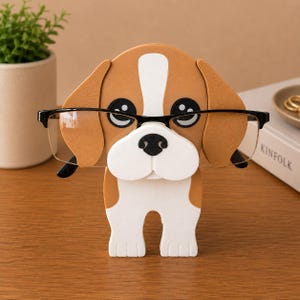 May include: A dog-shaped eyeglass holder in brown, white, and black, holding a pair of black-framed eyeglasses. The holder has large black eyes and a white snout. The eyeglasses are clear-lensed. The holder is made of a sturdy material.
