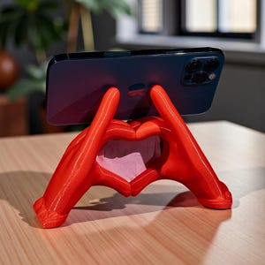 May include: A red phone stand shaped like hands forming a heart. The stand is holding a dark blue smartphone. The stand is on a light brown wooden surface.