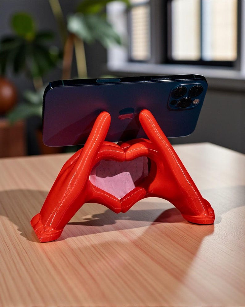 May include: A red phone stand shaped like hands forming a heart. The stand is holding a dark blue smartphone. The stand is on a light brown wooden surface.