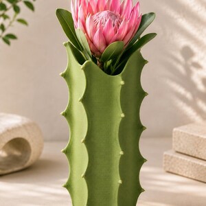 May include: A green cactus-shaped vase holds a vibrant pink protea flower with green leaves. The vase has a textured surface and small, light-colored protrusions along its sides. The overall aesthetic is modern and botanical.