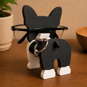 May include: A black and white dog-shaped eyeglasses holder with a key ring holder. The dog has large ears and a small tail. The eyeglasses are black-framed and rest on the dog's head. The key ring is silver.