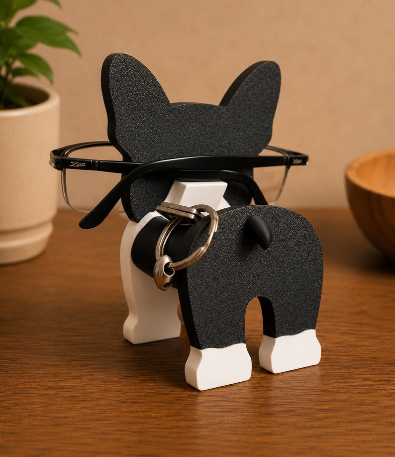 May include: A black and white dog-shaped eyeglasses holder with a key ring holder. The dog has large ears and a small tail. The eyeglasses are black-framed and rest on the dog's head. The key ring is silver.