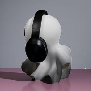 May include: A white bust sculpture with a black wireless headset resting on its head. The sculpture has a smooth, matte finish and is set against a pink surface and a gray background. The headphones are black and have a padded headband and earcups.