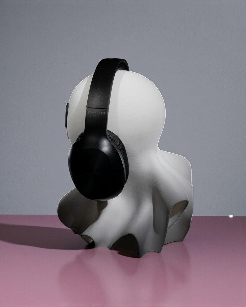 May include: A white bust sculpture with a black wireless headset resting on its head. The sculpture has a smooth, matte finish and is set against a pink surface and a gray background. The headphones are black and have a padded headband and earcups.