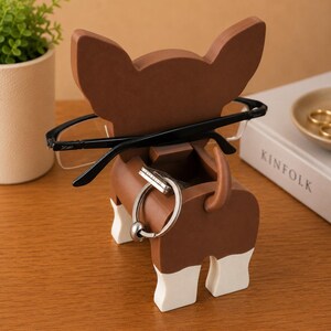 Puede incluir: Un soporte de madera marr&oacute;n y blanco para gafas y llaves con forma de perro. El soporte tiene una ranura para gafas y un anillo para llaves. Gafas negras descansan sobre el soporte. Una peque&ntilde;a planta y un libro est&aacute;n en el fondo.