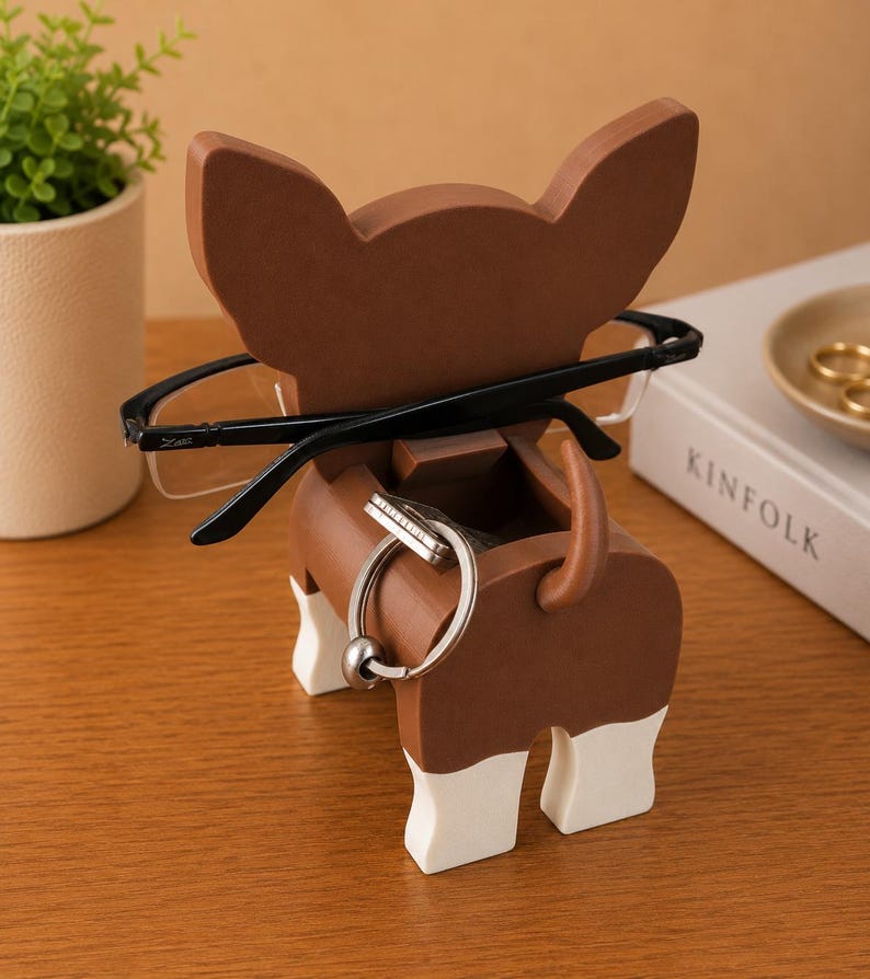 Puede incluir: Un soporte de madera marr&oacute;n y blanco para gafas y llaves con forma de perro. El soporte tiene una ranura para gafas y un anillo para llaves. Gafas negras descansan sobre el soporte. Una peque&ntilde;a planta y un libro est&aacute;n en el fondo.