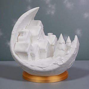 May include: A white ceramic crescent moon sculpture with a miniature village scene inside. The village features houses and trees, all in white, set on a gold-colored base. The background has a subtle snowflake pattern.