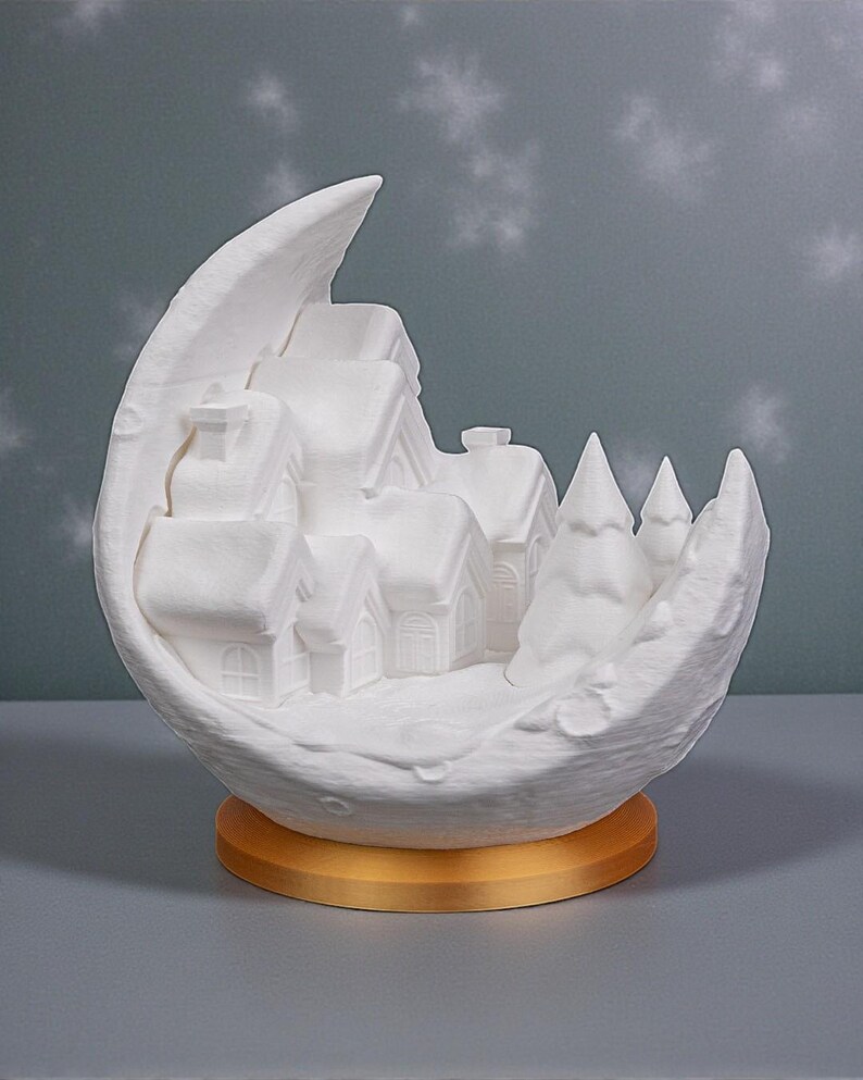 May include: A white ceramic crescent moon sculpture with a miniature village scene inside. The village features houses and trees, all in white, set on a gold-colored base. The background has a subtle snowflake pattern.
