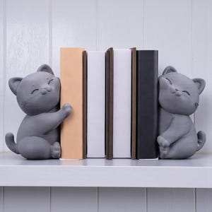 May include: Gray cat bookends with smiling faces, holding books on a white shelf. The books are in various colors, including beige, white, brown, and black. The bookends are a cute and functional addition to any bookshelf.