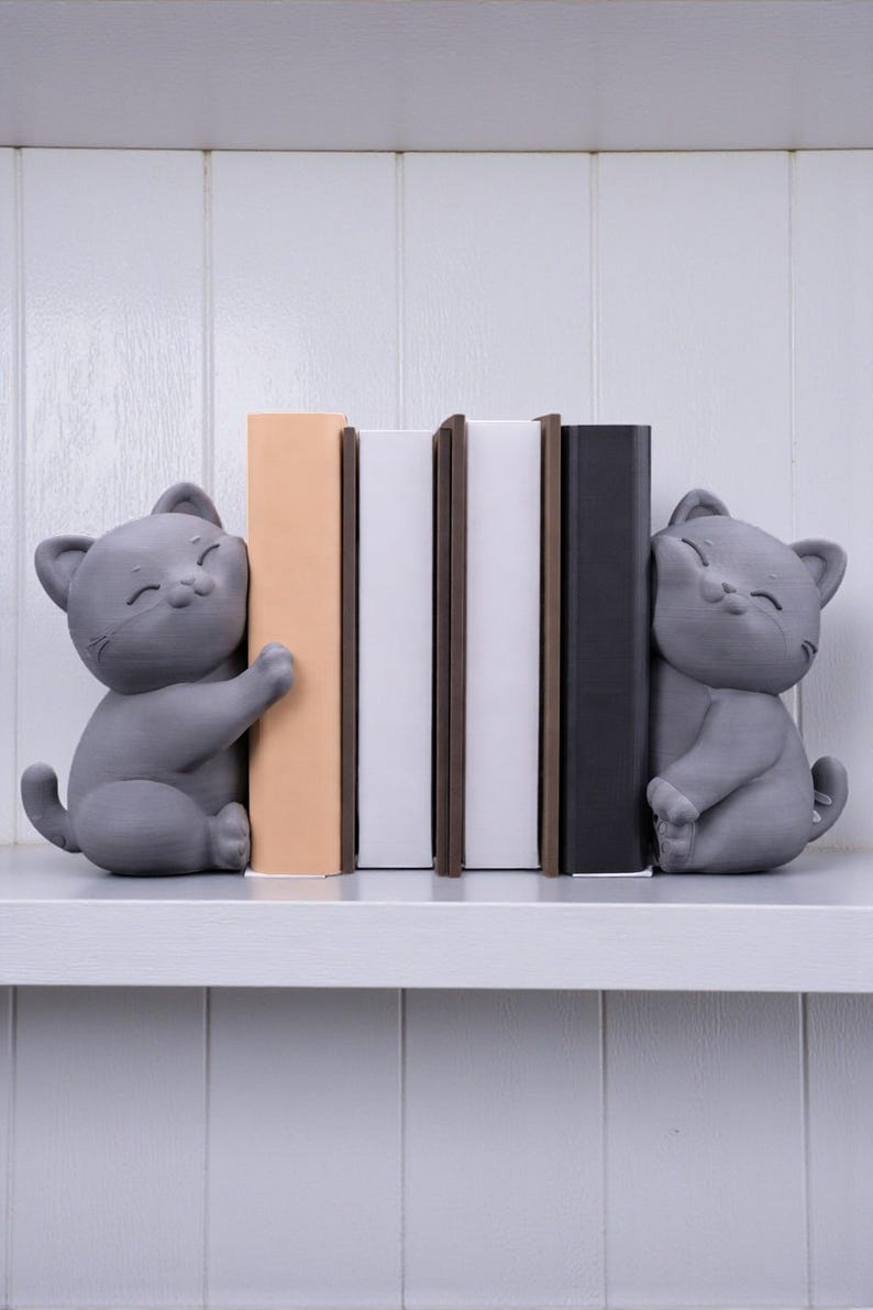 May include: Gray cat bookends with smiling faces, holding books on a white shelf. The books are in various colors, including beige, white, brown, and black. The bookends are a cute and functional addition to any bookshelf.