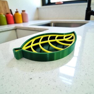 May include: A green and yellow leaf-shaped soap dish. The dish has a dark green base with a yellow, leaf-vein design on top. The dish is made of plastic and sits on a white countertop. The dish is designed to hold a bar of soap.