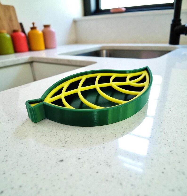 May include: A green and yellow leaf-shaped soap dish. The dish has a dark green base with a yellow, leaf-vein design on top. The dish is made of plastic and sits on a white countertop. The dish is designed to hold a bar of soap.