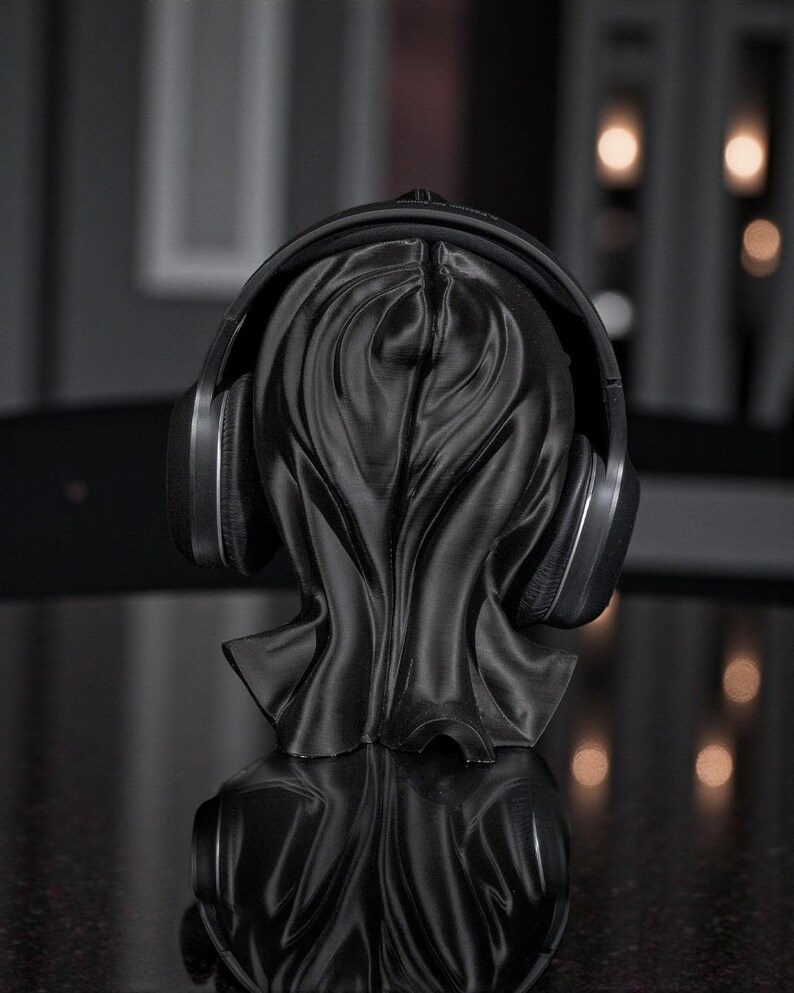 May include: A black headphone stand shaped like a draped figure, holding a pair of black and silver headphones. The stand has a textured surface and is reflected on a dark, glossy surface. The background is blurred with soft lighting.