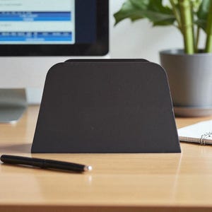 K&ouml;nnte beinhalten: Ein schwarzer Schreibtisch-Organizer mit trapezf&ouml;rmiger Form, der auf einer hellen Holzoberfl&auml;che steht. Ein schwarzer Stift liegt vor dem Organizer. Ein Computermonitor und eine Topfpflanze befinden sich im Hintergrund.