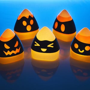 May include: Five Halloween-themed candy corn shaped decorations with different facial expressions. The decorations are lit from within, casting a warm glow.