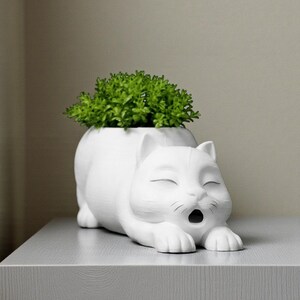 May include: A white ceramic cat-shaped planter with a green plant inside. The cat is lying down with its eyes closed and mouth open. The planter is on a gray surface.