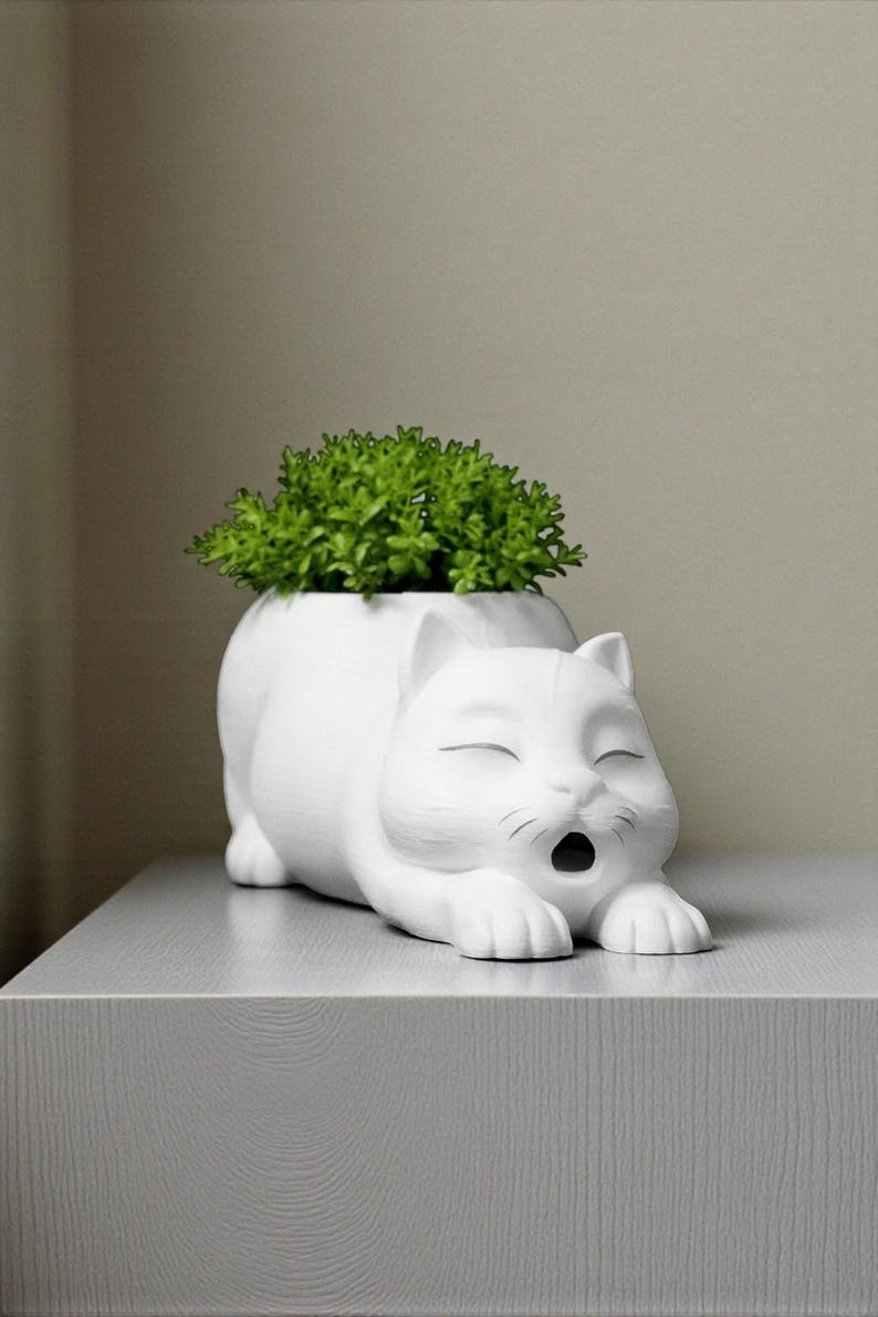 May include: A white ceramic cat-shaped planter with a green plant inside. The cat is lying down with its eyes closed and mouth open. The planter is on a gray surface.