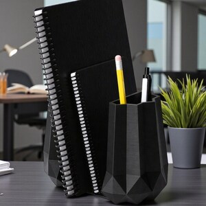 May include: A black desk organizer with a geometric design holds a yellow pencil, a black pen, and a white pen. Two black spiral notebooks are behind the organizer. A small potted plant is to the right.