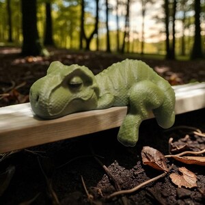 May include: A light green T-Rex figurine resting on a wooden plank. The dinosaur has detailed skin texture and is lying down with its head and one arm hanging over the edge. The background is a blurred forest scene.