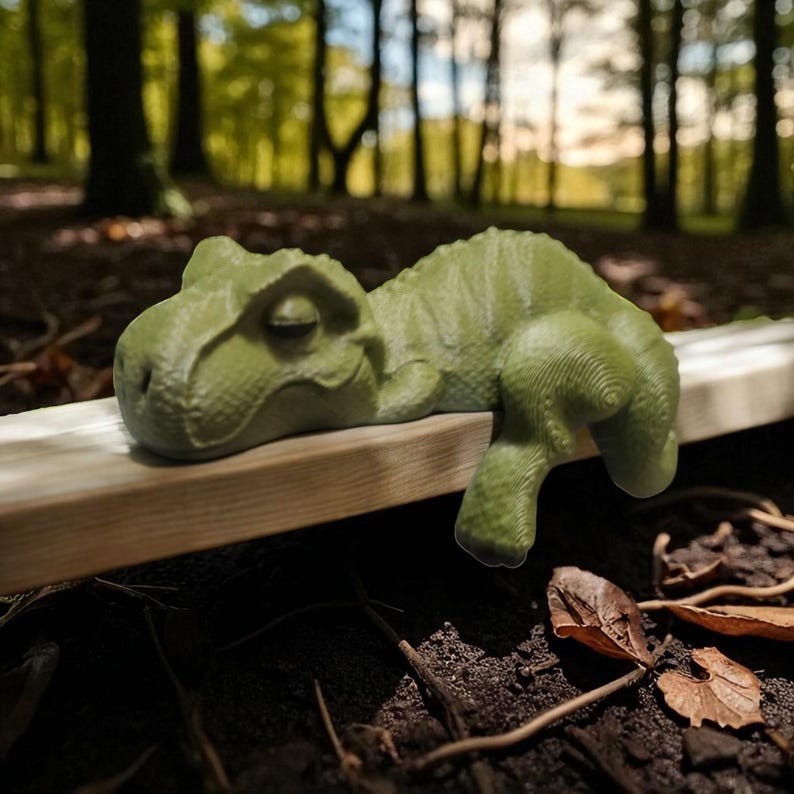 May include: A light green T-Rex figurine resting on a wooden plank. The dinosaur has detailed skin texture and is lying down with its head and one arm hanging over the edge. The background is a blurred forest scene.