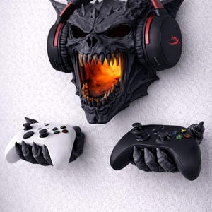 May include: A black and gray demon skull wall mount with glowing orange light in the mouth, holding a black headset with red accents. Two game controllers, one white and one black, are held by claw-like hands.