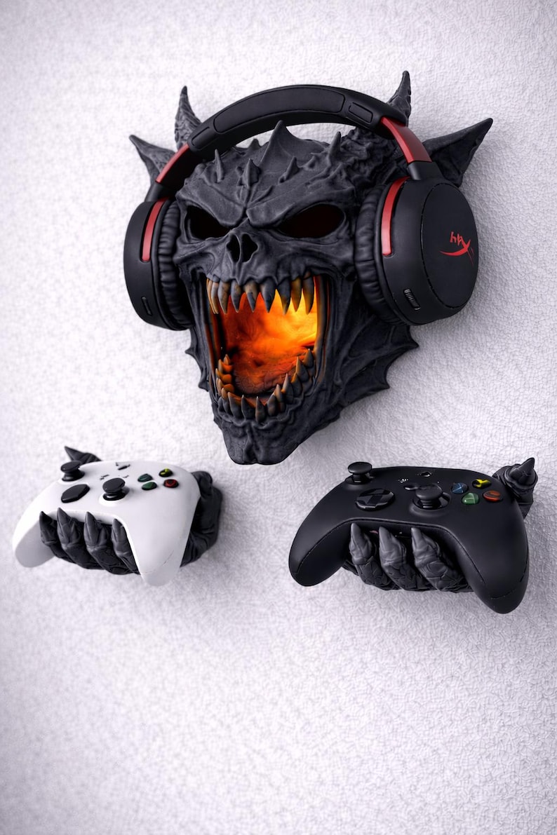 May include: A black and gray demon skull wall mount with glowing orange light in the mouth, holding a black headset with red accents. Two game controllers, one white and one black, are held by claw-like hands.