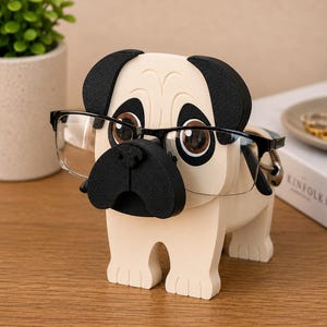 May include: A beige pug-shaped eyeglass holder with black ears, nose, and a pair of black-framed glasses. The holder has brown eyes and is standing on a wooden surface. A small green plant in a white pot is in the background.