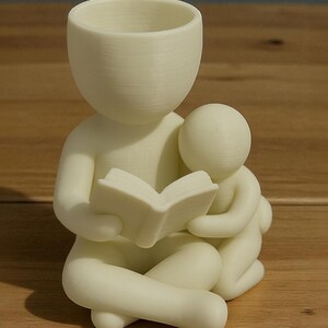 May include: A cream-colored figurine depicts a person reading a book to a smaller figure. The larger figure has a hollow head, and the smaller figure is nestled close. The figures are seated with their legs crossed. The figurine is on a wooden surface.