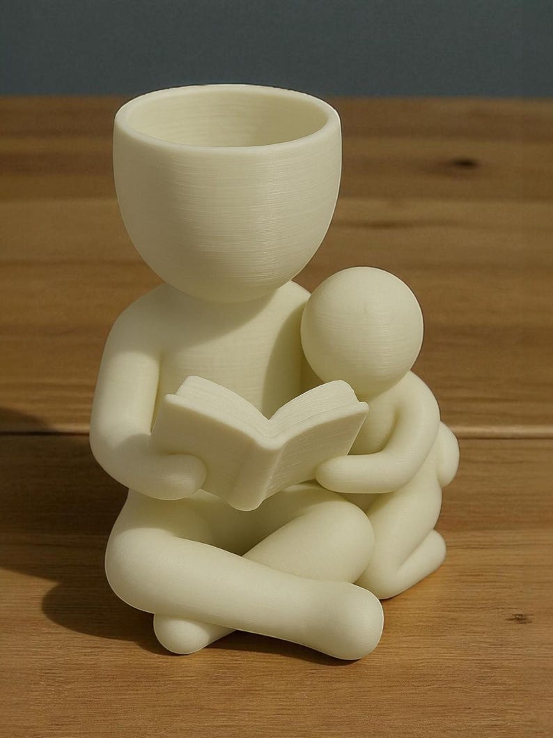 May include: A cream-colored figurine depicts a person reading a book to a smaller figure. The larger figure has a hollow head, and the smaller figure is nestled close. The figures are seated with their legs crossed. The figurine is on a wooden surface.