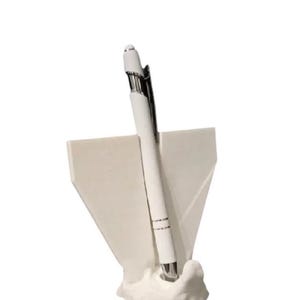 May include: A white pen with silver accents is displayed in a white pen holder. The pen holder has a unique, abstract design. The pen is angled slightly, showcasing its sleek, modern aesthetic.