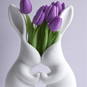 May include: A white ceramic bunny vase holding a bouquet of purple tulips. The vase is shaped like two bunnies embracing, with the flowers emerging from their joined bodies. The tulips have green stems and leaves.