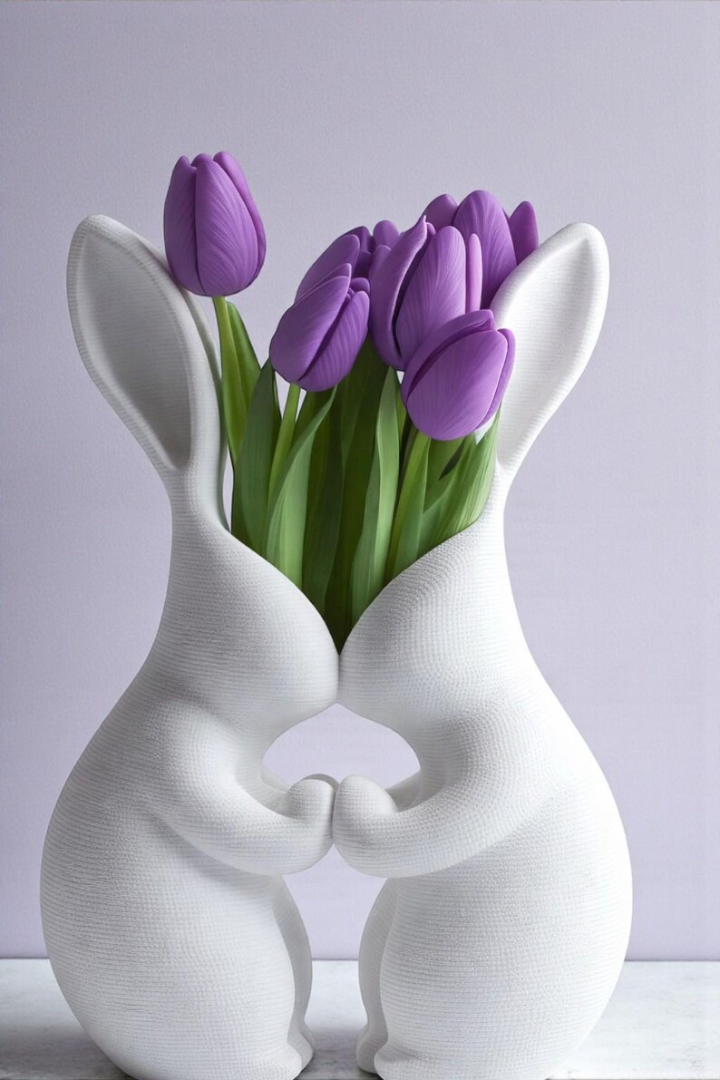 May include: A white ceramic bunny vase holding a bouquet of purple tulips. The vase is shaped like two bunnies embracing, with the flowers emerging from their joined bodies. The tulips have green stems and leaves.