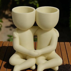 May include: A cream-coloured ceramic planter shaped like two figures embracing. Each figure has a rounded head with a hollowed-out top for planting. The figures are seated with their legs crossed and hands clasped, creating a unique decorative piece.